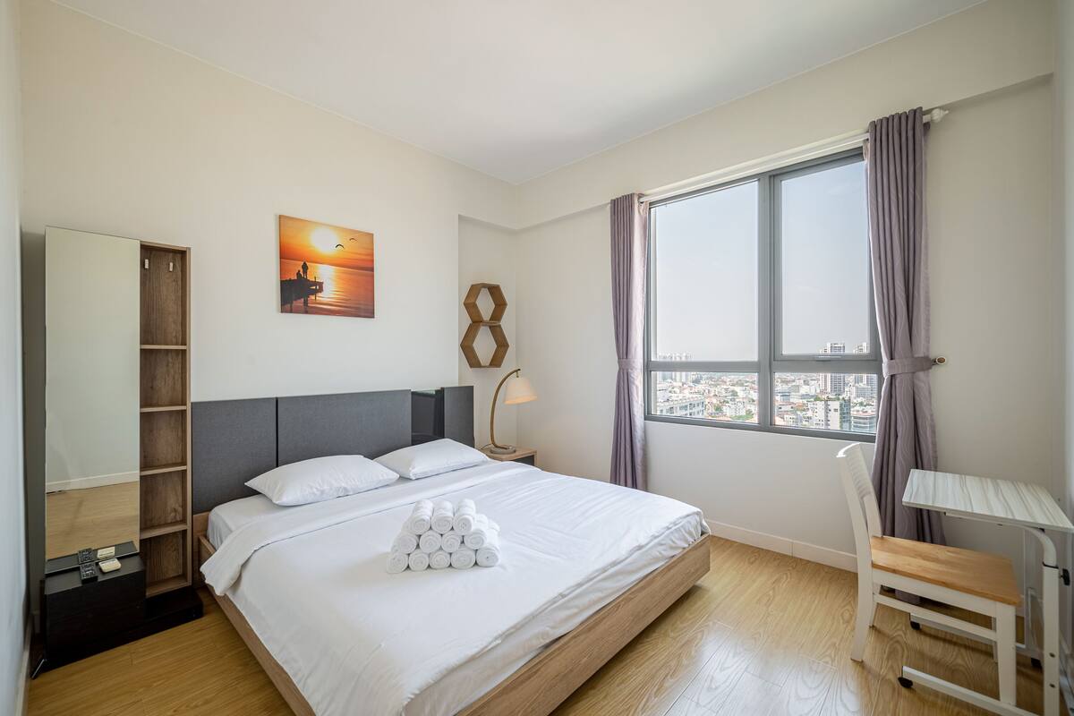 A hotel room with a bed, a desk, a chair, a window with a city view, a painting, and a wooden wardrobe.