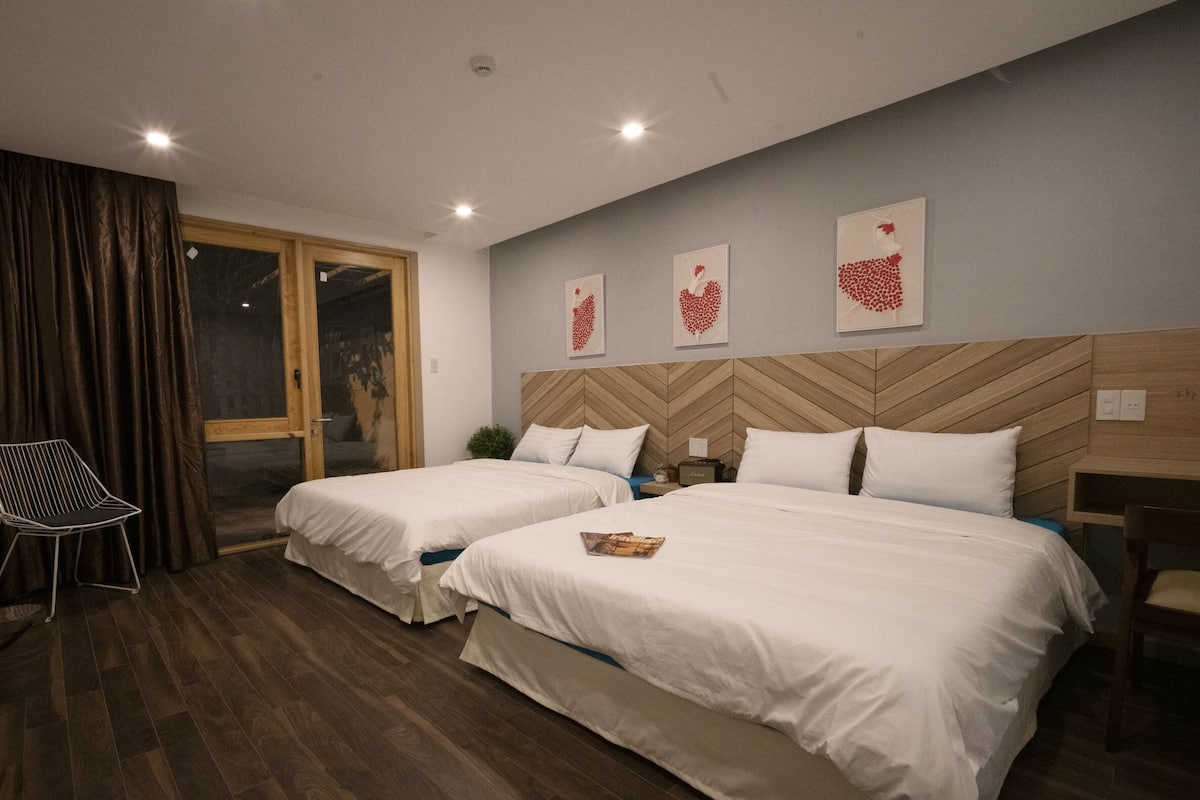 A hotel room with two beds, a wooden headboard, a desk, a chair, and a window with curtains.