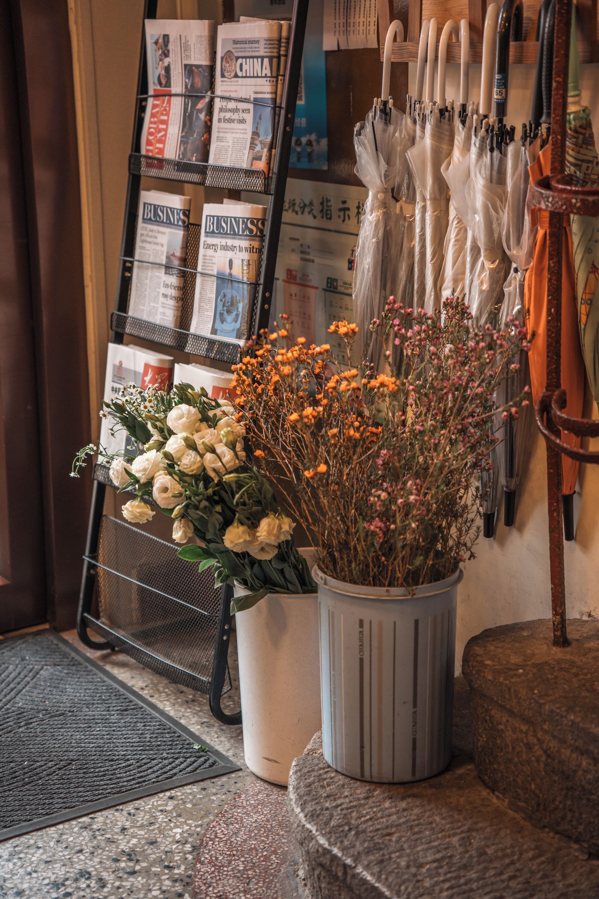Un arreglo floral con flores blancas y naranjas, un revistero con revistas de negocios, y varios paraguas colgados en un perchero.