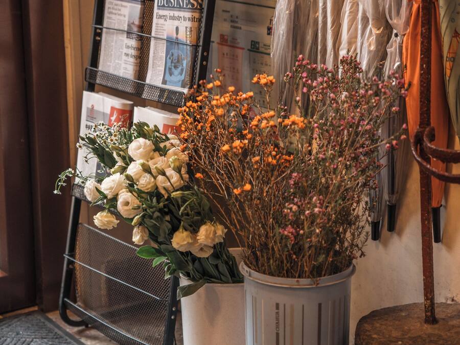 Un arreglo floral con flores blancas y naranjas, un revistero con revistas de negocios, y varios paraguas colgados en un perchero.