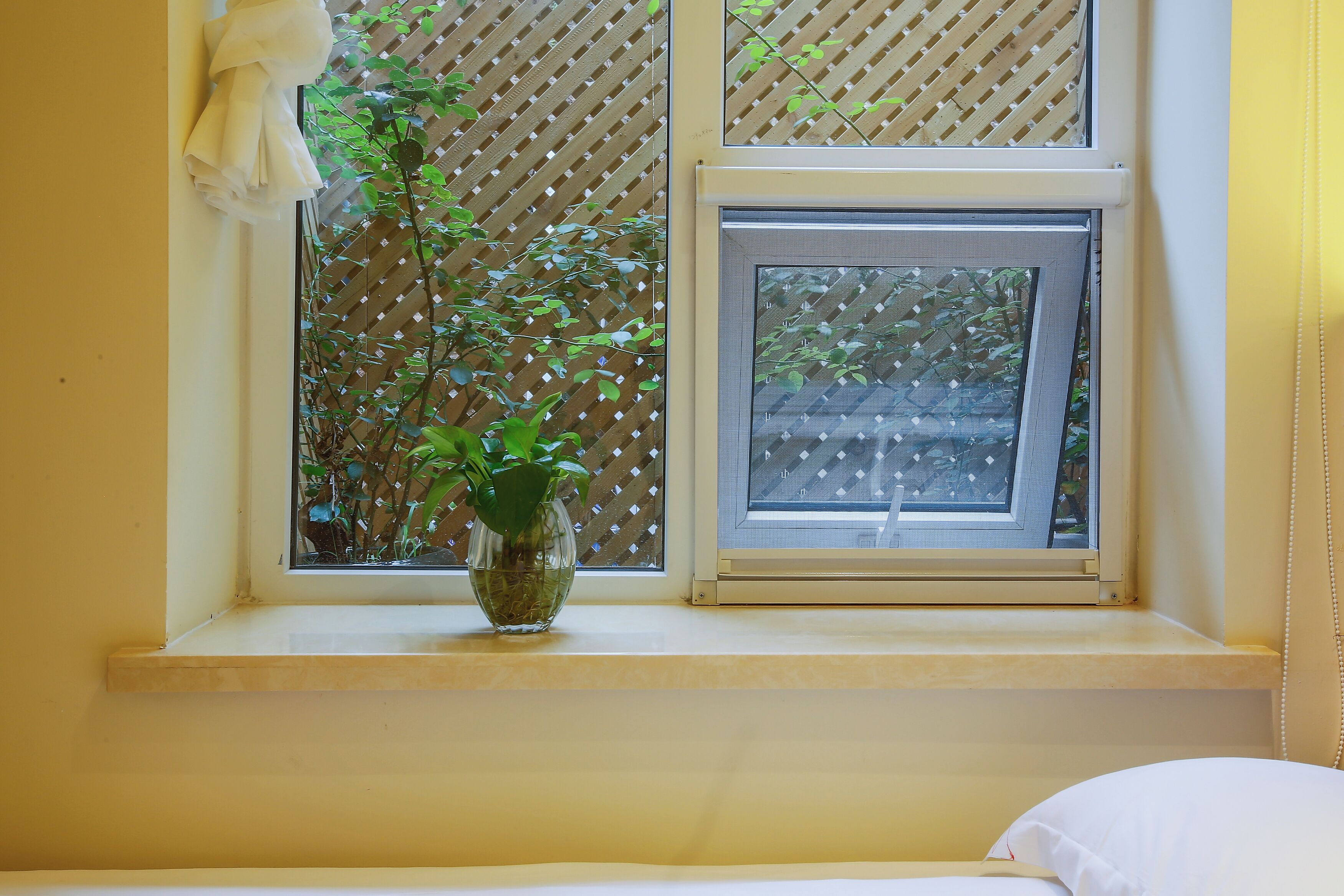A window with a plant in a vase, a bed with white bedding, and a yellow wall.