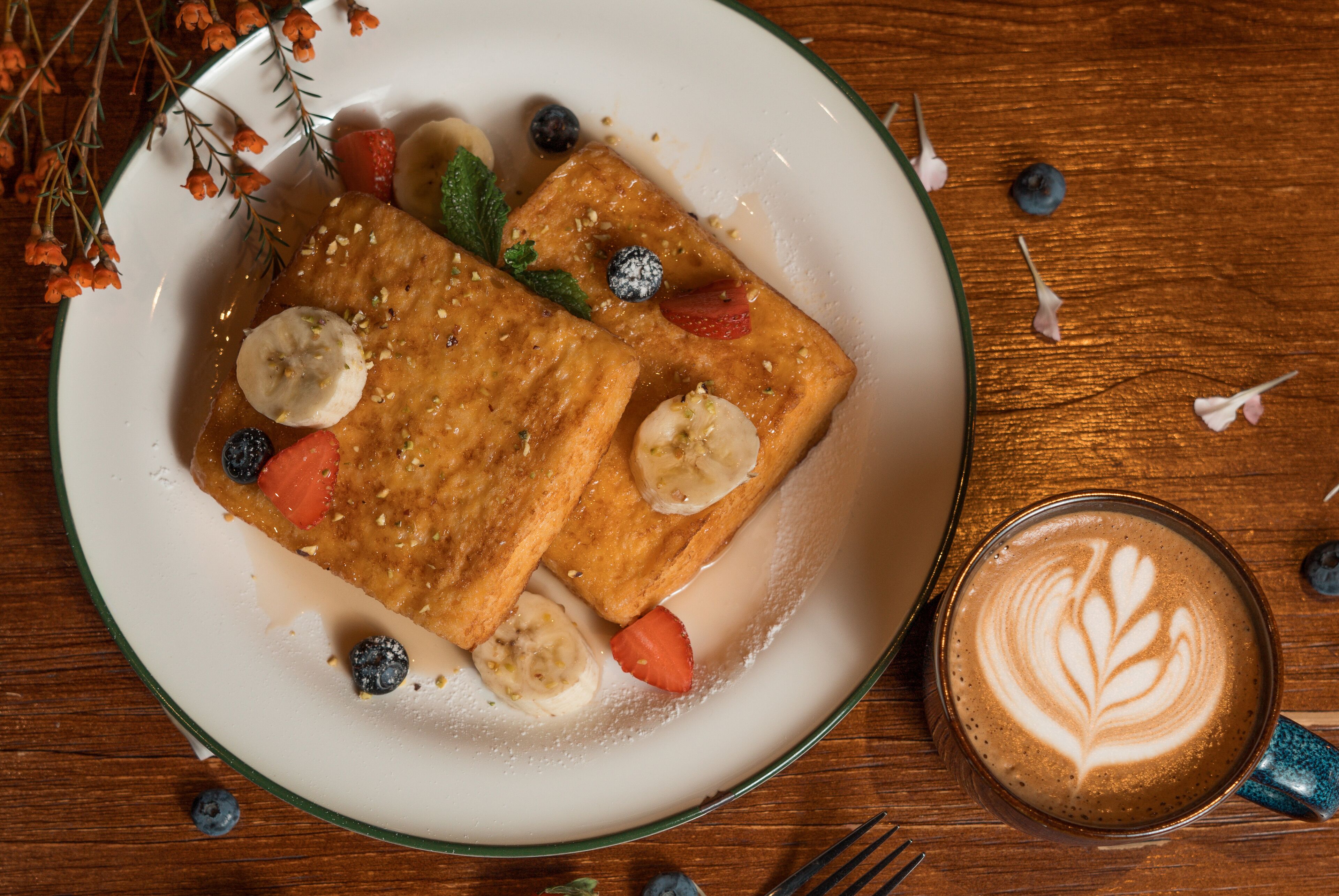 A plate of pancakes with fruit and syrup, and a cup of coffee with latte art on a wooden table.