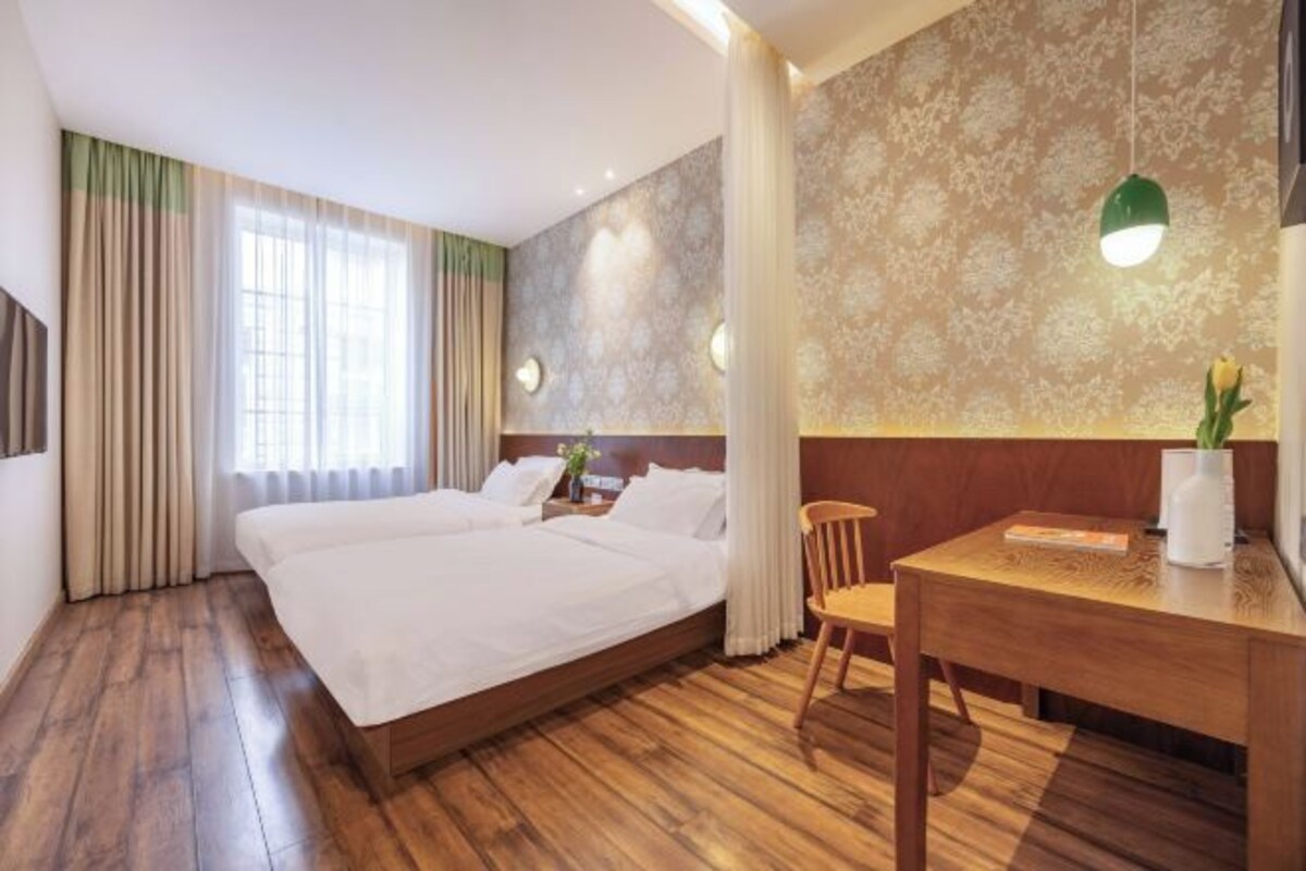 A hotel room with a large bed, a wooden desk, and a chair. There is a window with curtains, a wall-mounted lamp, and a vase with a plant.