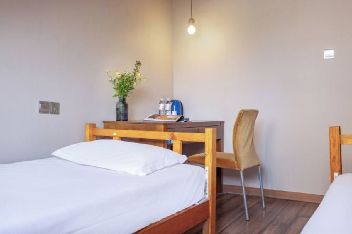 A bedroom with a wooden bed, a desk with a chair, a vase with flowers, and a hanging light bulb.
