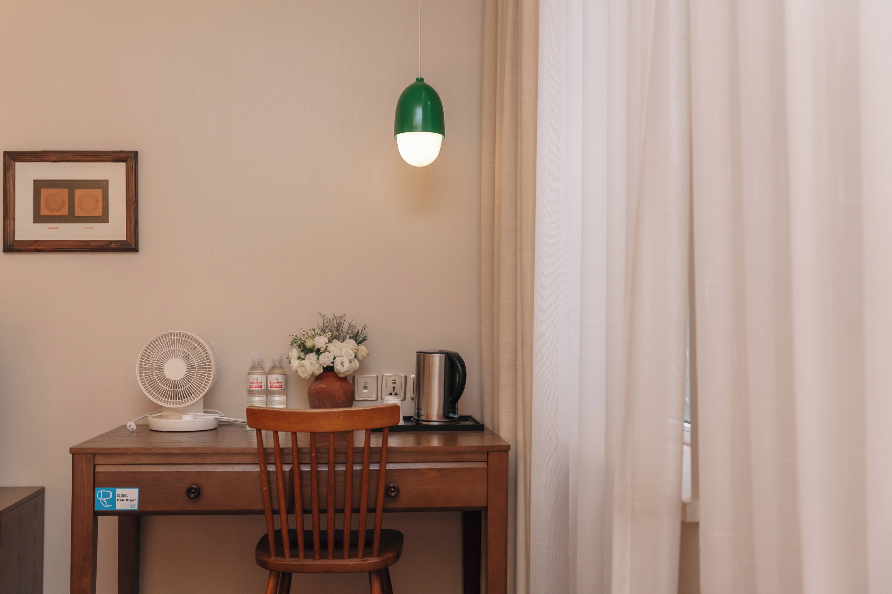 Un escritorio de madera con una silla, una lámpara colgante verde, un cuadro enmarcado, un ventilador, un jarrón con flores, una tetera y un enchufe.
