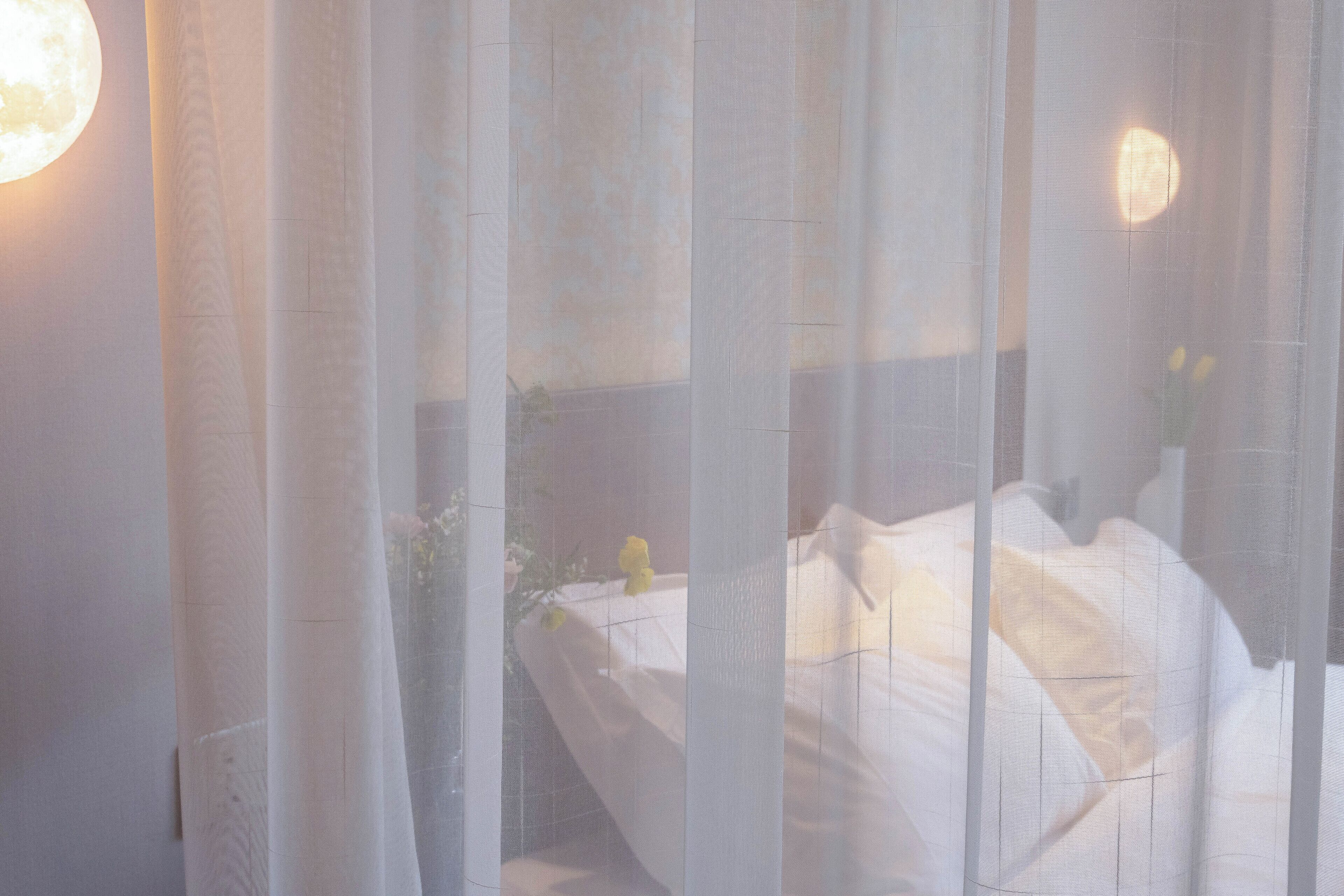 A room with a bed covered in white linens, a nightstand with a lamp, and a window with sheer curtains.