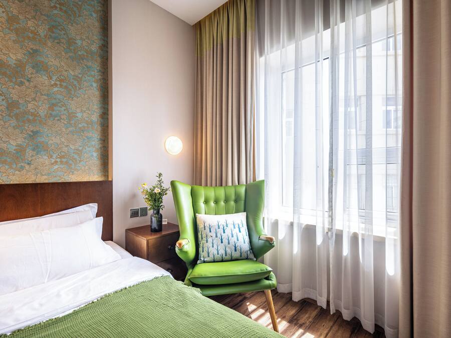A hotel room with a bed, a green armchair with a patterned pillow, a nightstand with a vase of flowers, and a large window with sheer curtains.