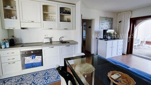 Une cuisine moderne avec des armoires blanches, un four encastré et un évier. On y trouve un coin repas avec une table et des chaises en verre, et une vue sur l’extérieur grâce à une porte vitrée.