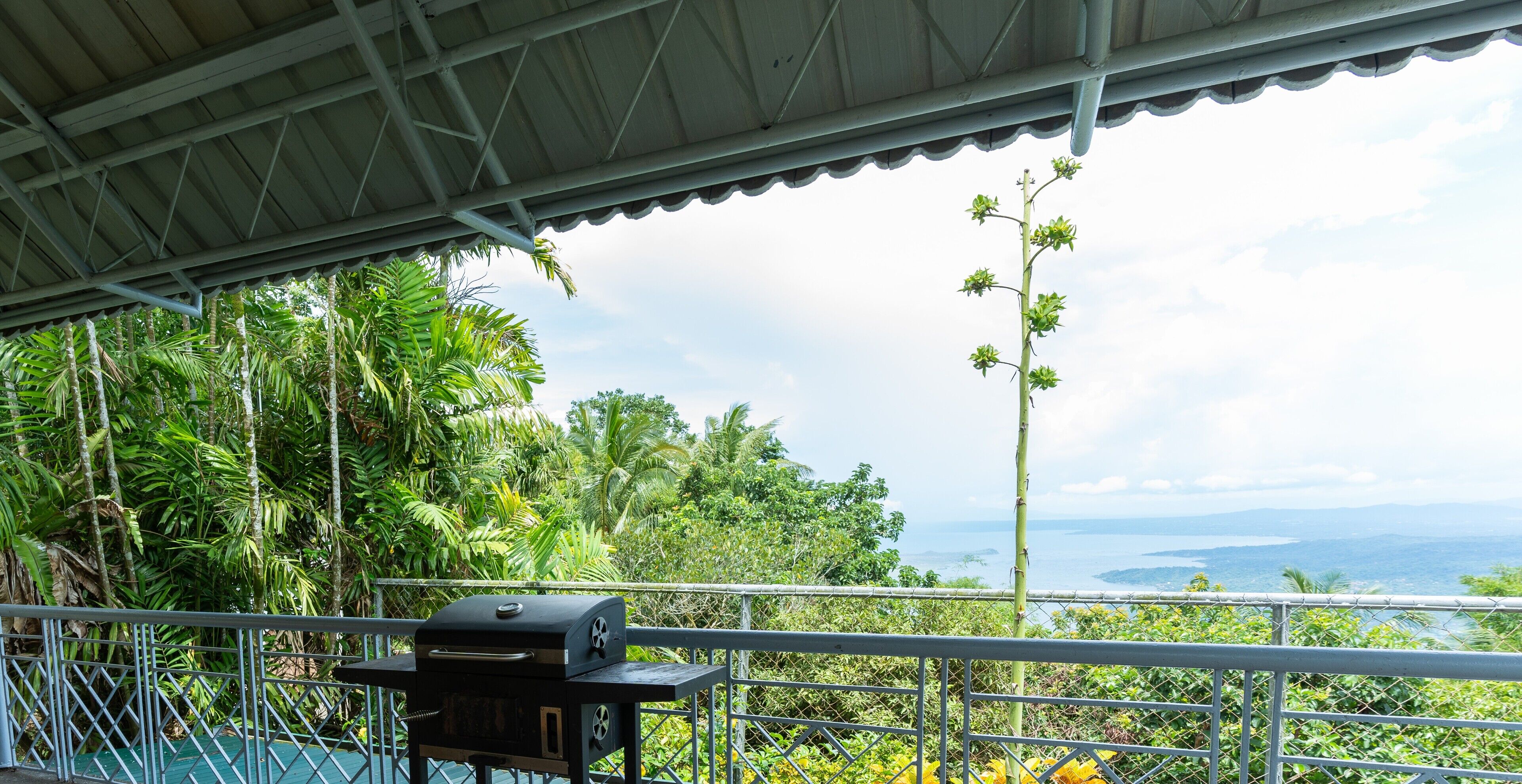 A covered outdoor area with a grill, surrounded by lush greenery and a scenic view of the ocean.