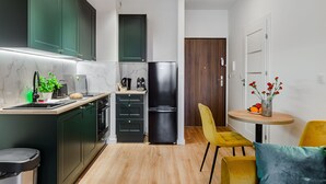 Een kleine keuken met donkere kastjes, een zwarte koelkast, een spoelbak en een eethoekje met een gele stoel en een tafel.