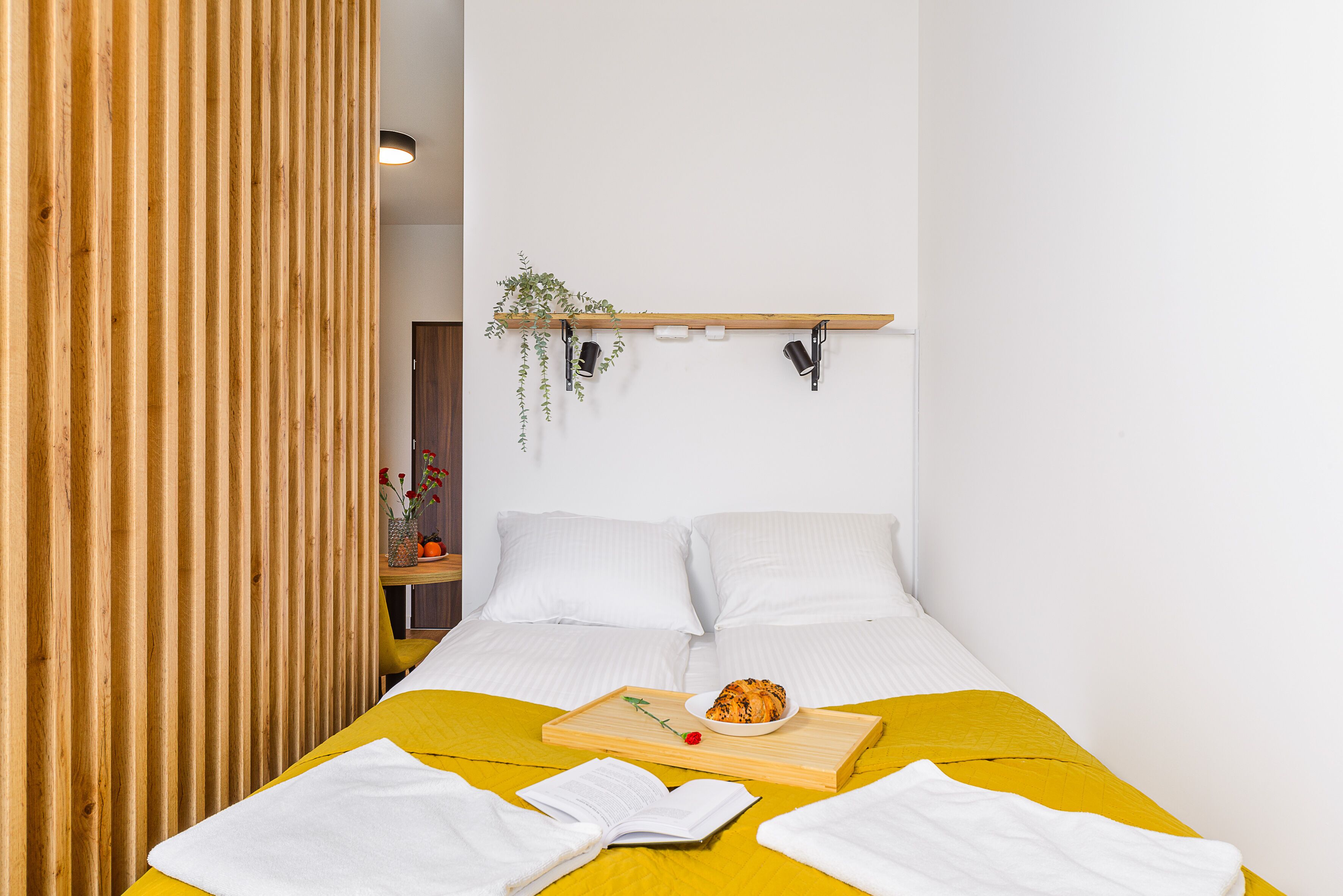 A room with a bed, a bedside table, a shelf with plants, and a tray with a book and a flower.