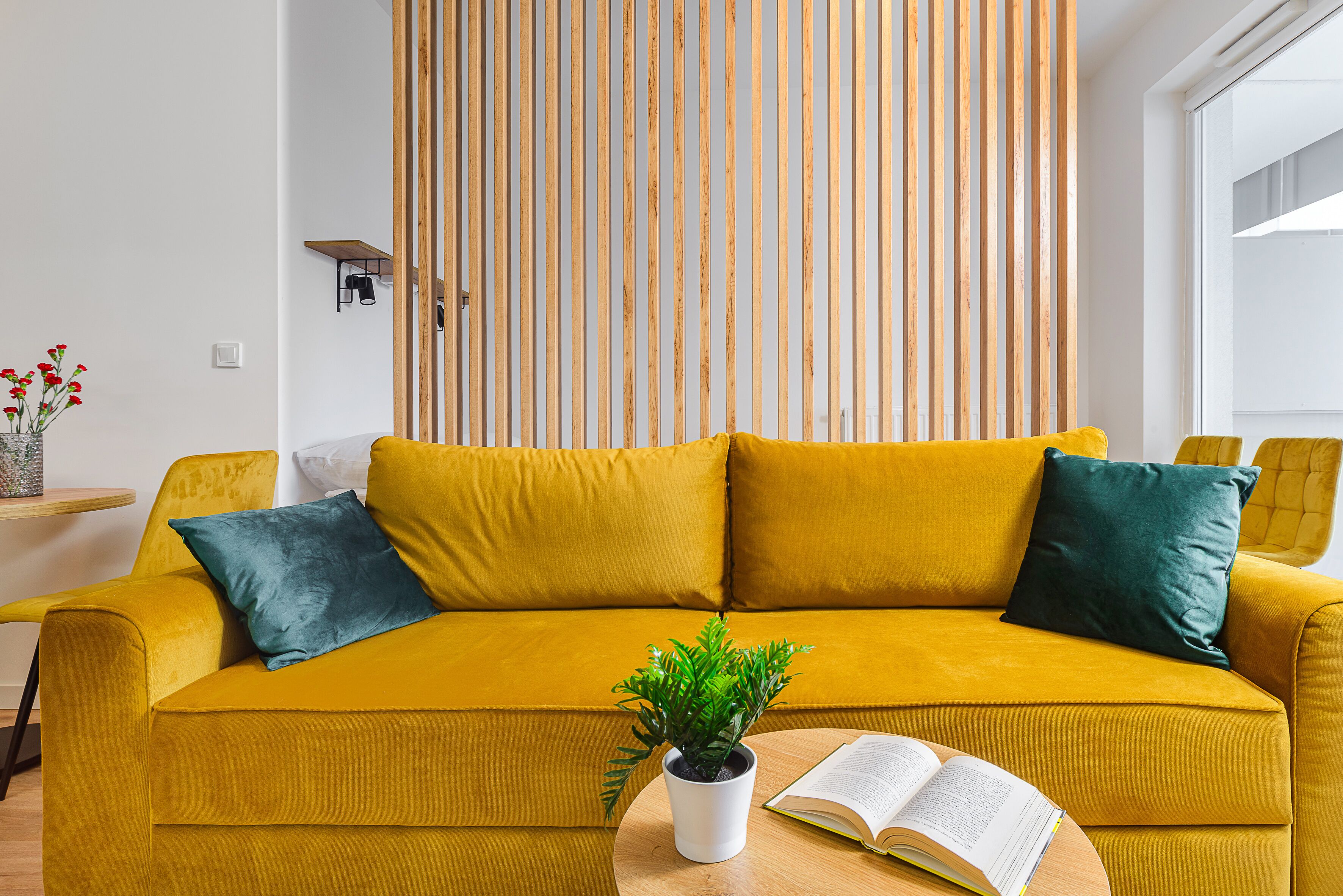 A mustard-colored sofa with two pillows, a potted plant, and an open book on a round wooden table.