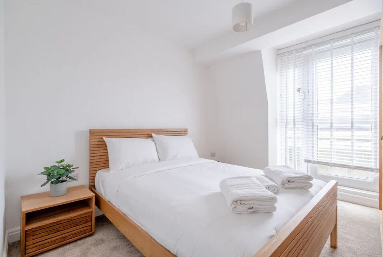 A neatly made bed with white linens and two folded towels, a bedside plant, and a window with blinds.