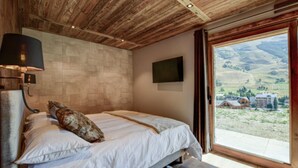 Une chambre avec un plafond en bois, un lit avec une literie blanche, un téléviseur fixé au mur et une grande fenêtre donnant sur des bâtiments et des collines.