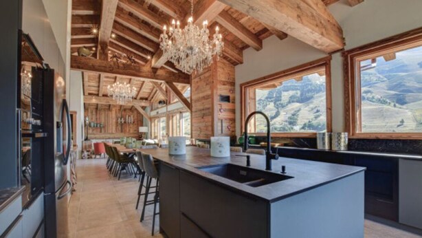 Une cuisine moderne dotée d’un îlot central, de poutres en bois et de grandes fenêtres offrant une vue sur les montagnes.