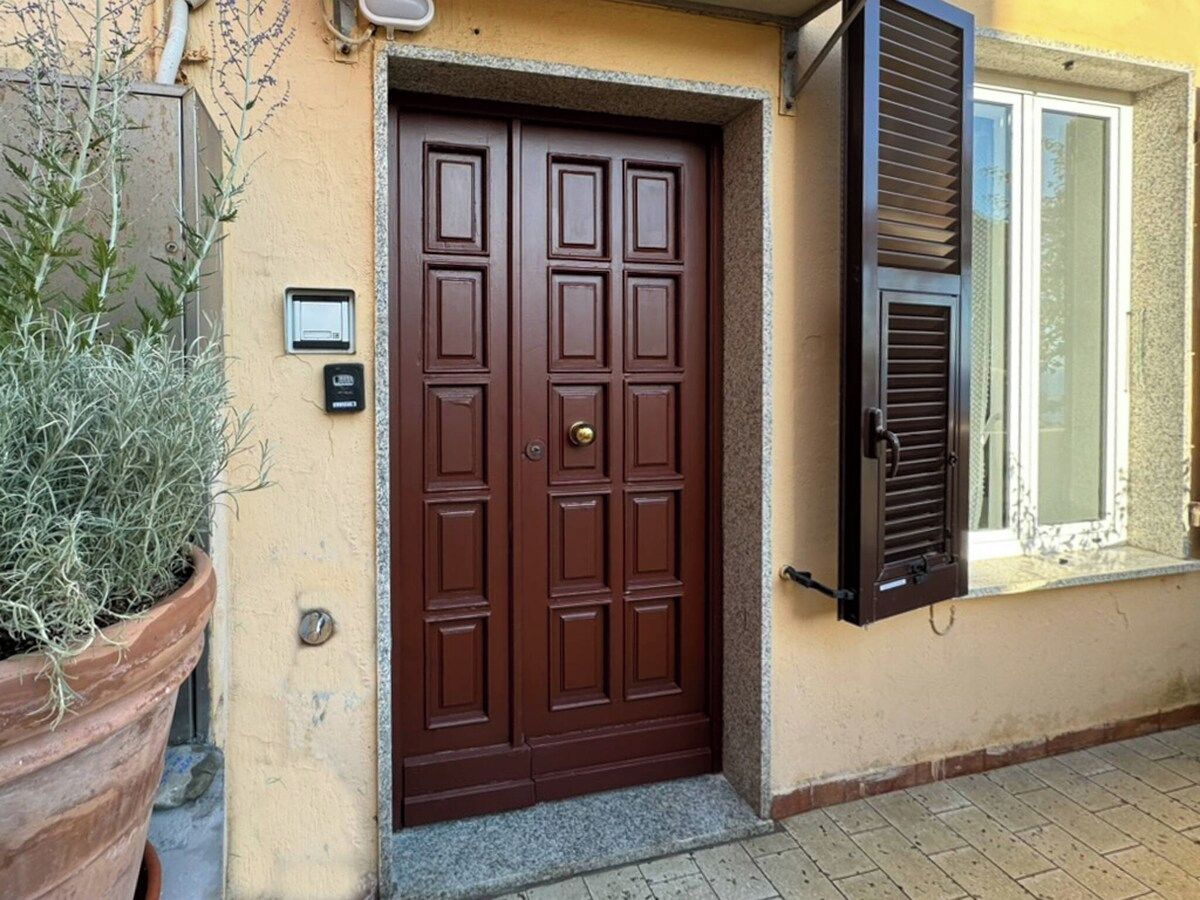Eine braune Tür mit einem Schlüsselfeld, eine Topfpflanze links und ein Fenster mit Fensterläden rechts.