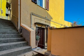 Un escalier étroit menant à une porte surmontée d’un portail en métal, un mur jaune et un ciel d’un bleu limpide.