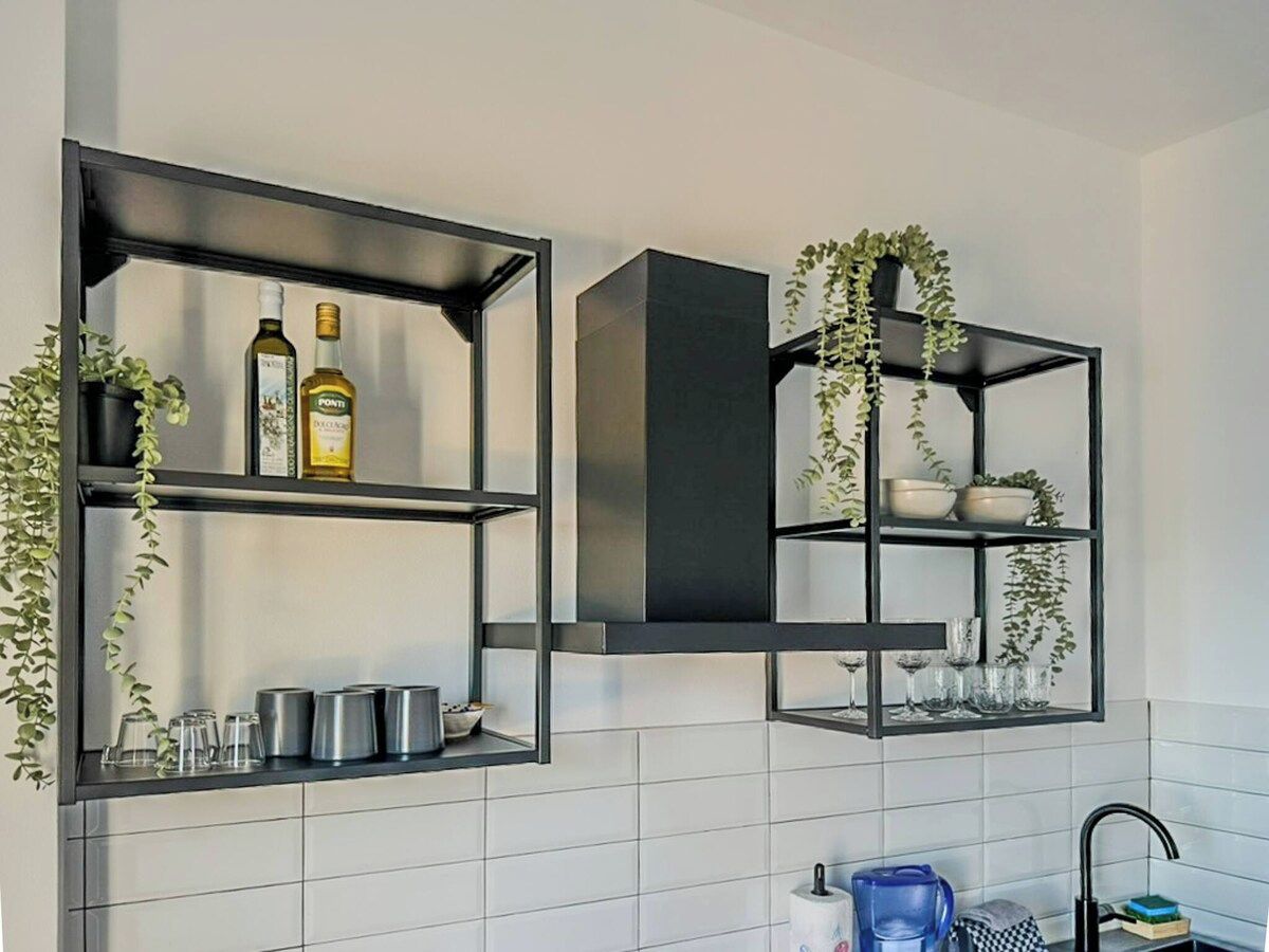 Une cuisine dotée d’étagères en métal noir, d’un dosseret de carrelage blanc et de plantes suspendues.