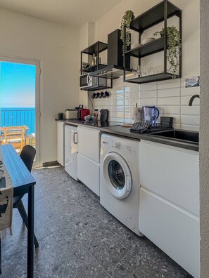A compact kitchen with a built-in washing machine, a dining area with a table and chairs, and a view of the sea through an open door. - Il Sogno di Beatrice Luxury Apartment (Manarola)