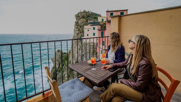Dos personas sentadas en una mesa con bebidas, con vistas a la costa y edificios.
