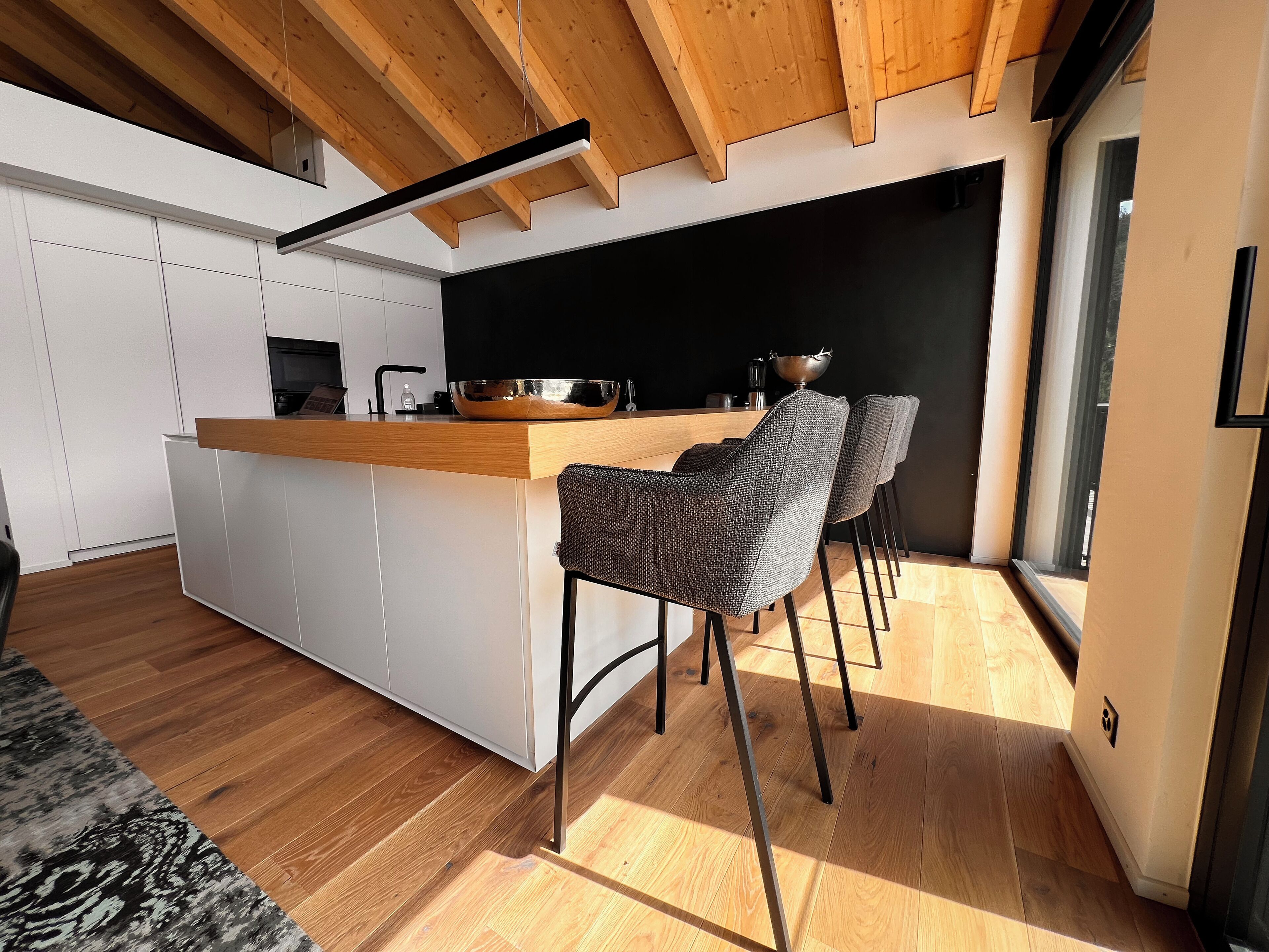 A modern kitchen with a wooden ceiling, white cabinets, and a central island. There are four bar stools and a large window providing natural light.