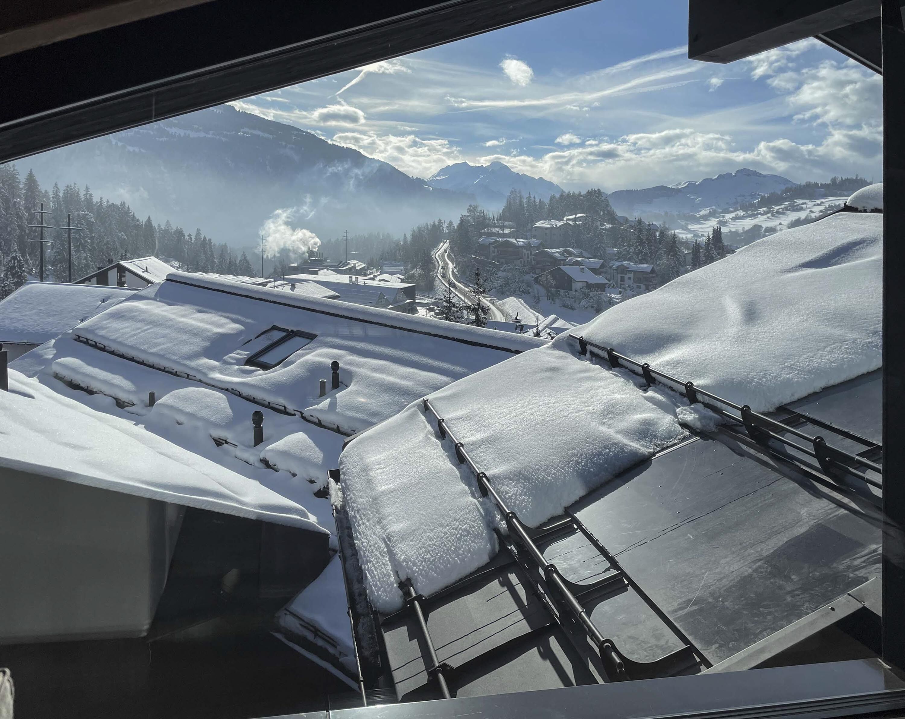 A snowy mountain landscape with a clear sky, visible through a window overlooking a snow-covered rooftop.
