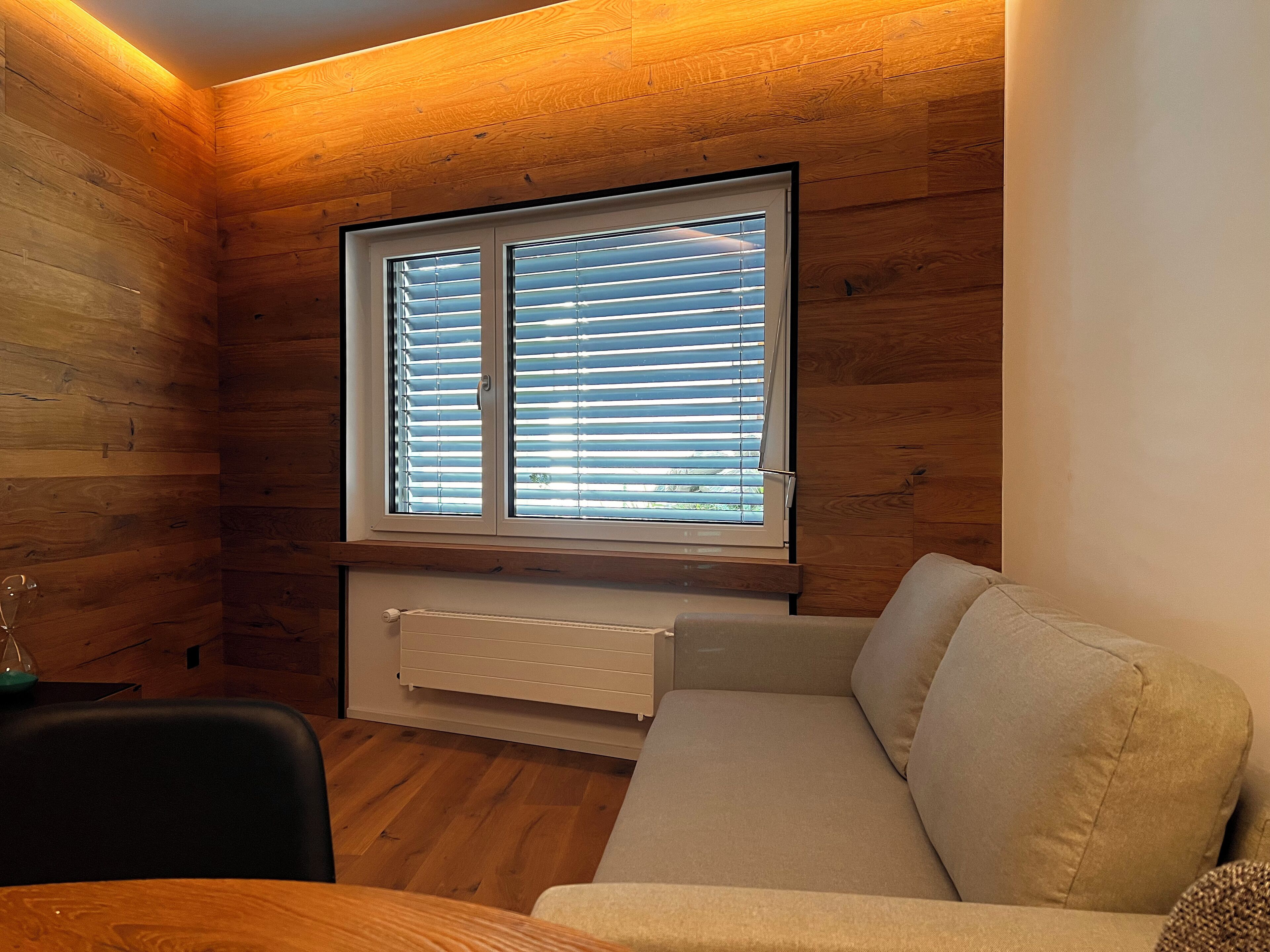 A cozy room with wooden walls, a window with blinds, a radiator, and a beige sofa.