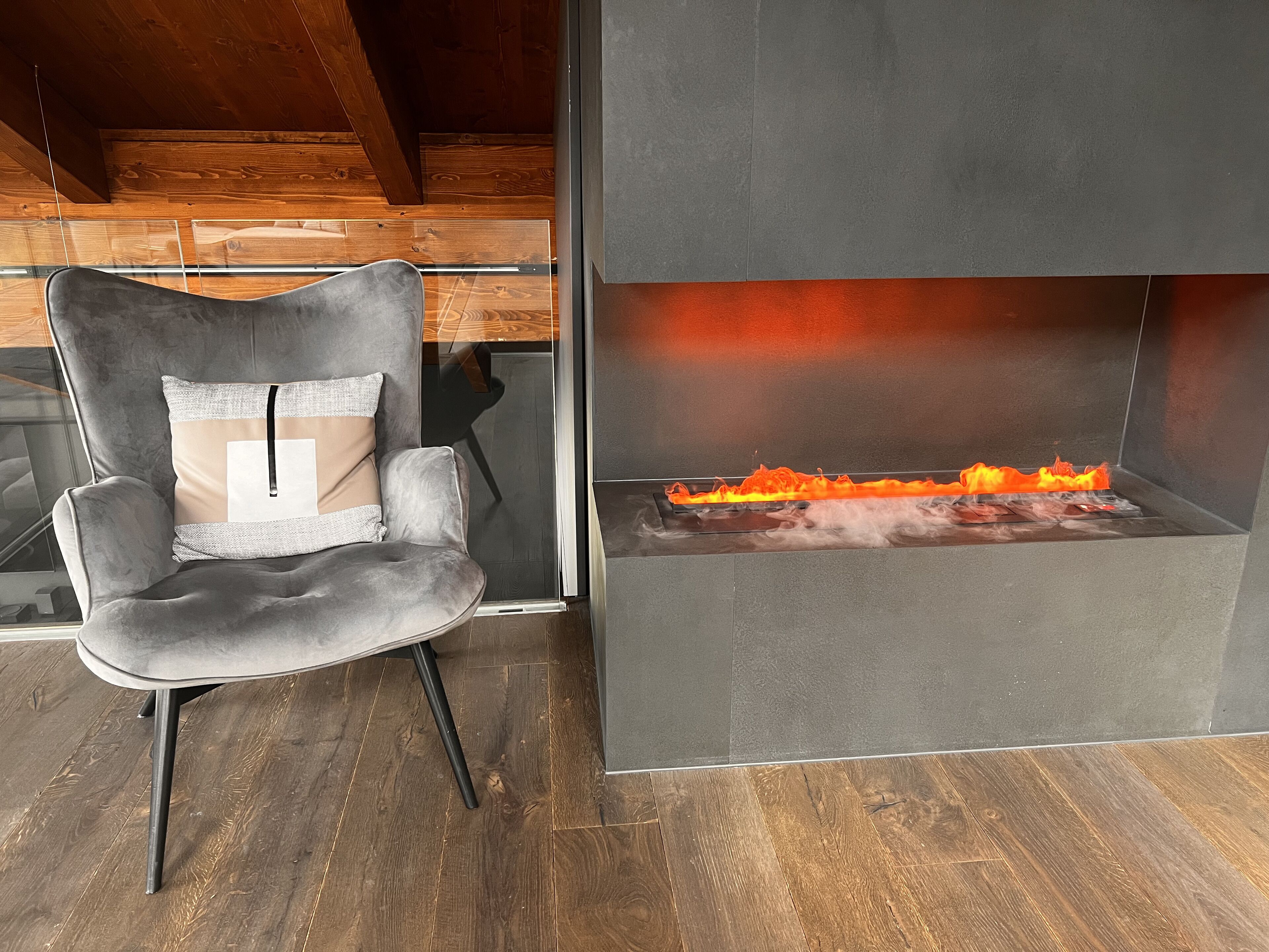 Una habitación moderna con una butaca tapizada de gris, una chimenea con fuego visible y suelo de madera.