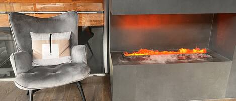 Una habitación moderna con una butaca tapizada de gris, una chimenea con fuego visible y suelo de madera.