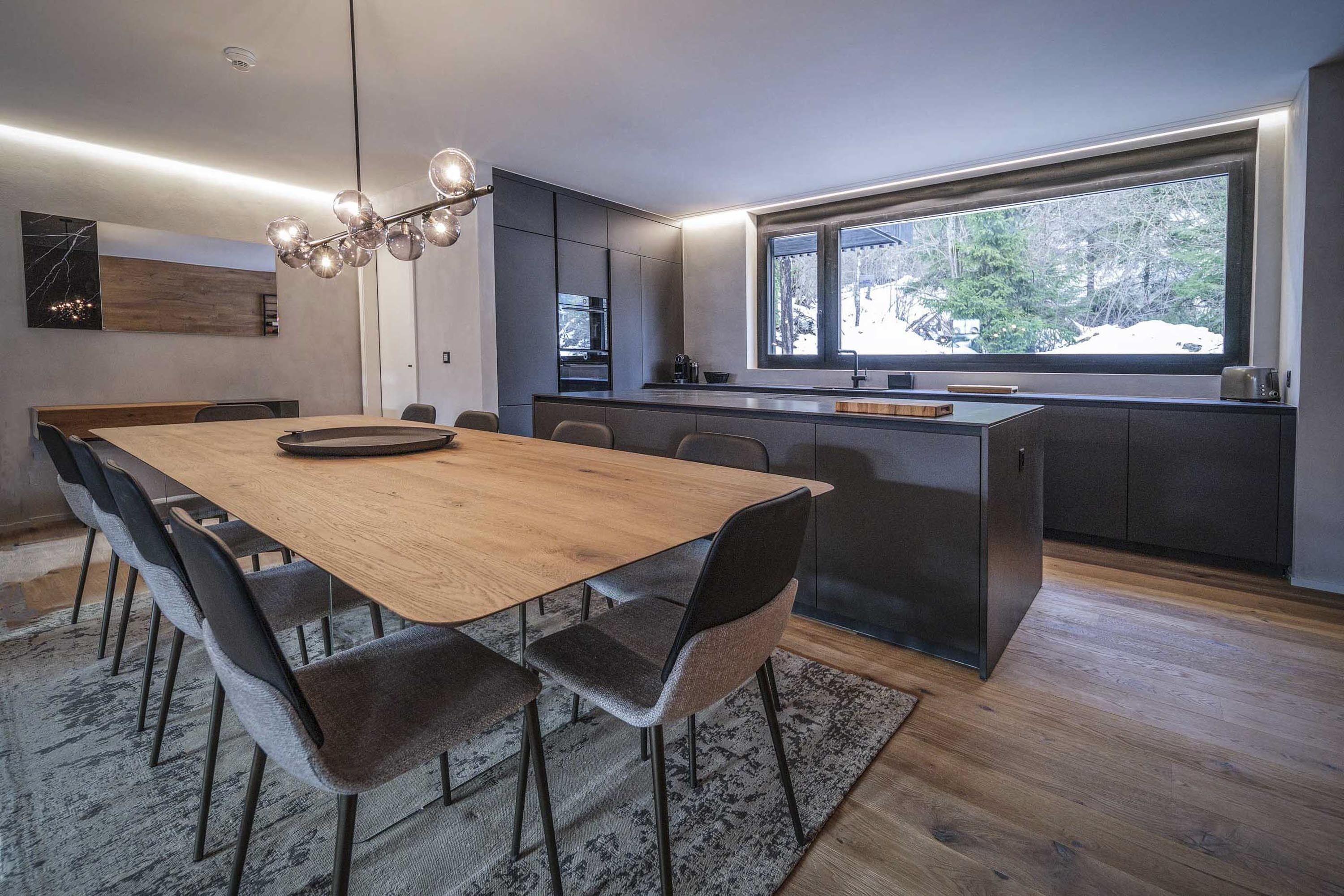 A modern kitchen with a wooden dining table and chairs, a built-in island, and a large window with a view of trees.