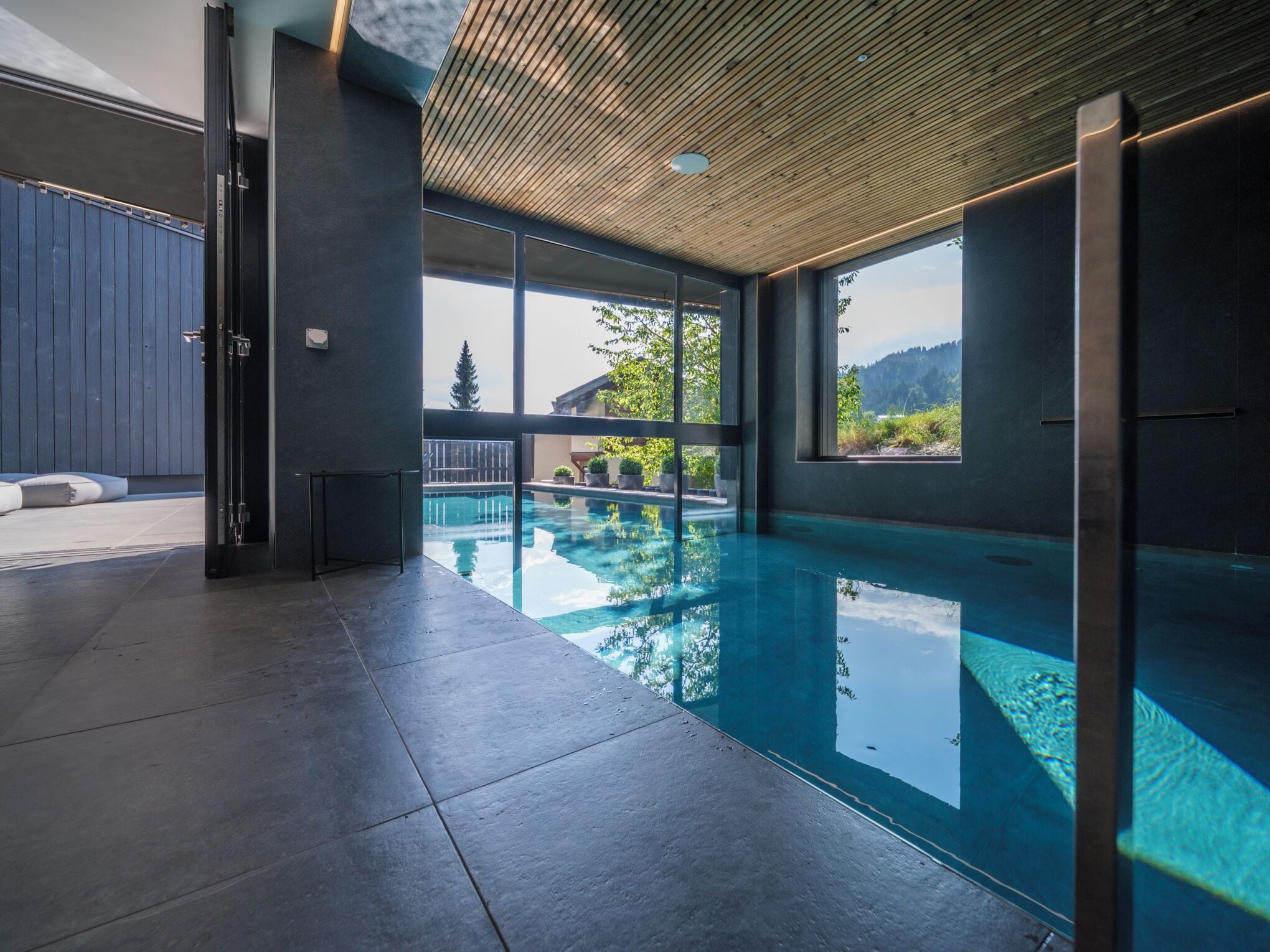 A modern indoor swimming pool with clear glass walls, a wooden ceiling, and a view of a mountainous landscape.
