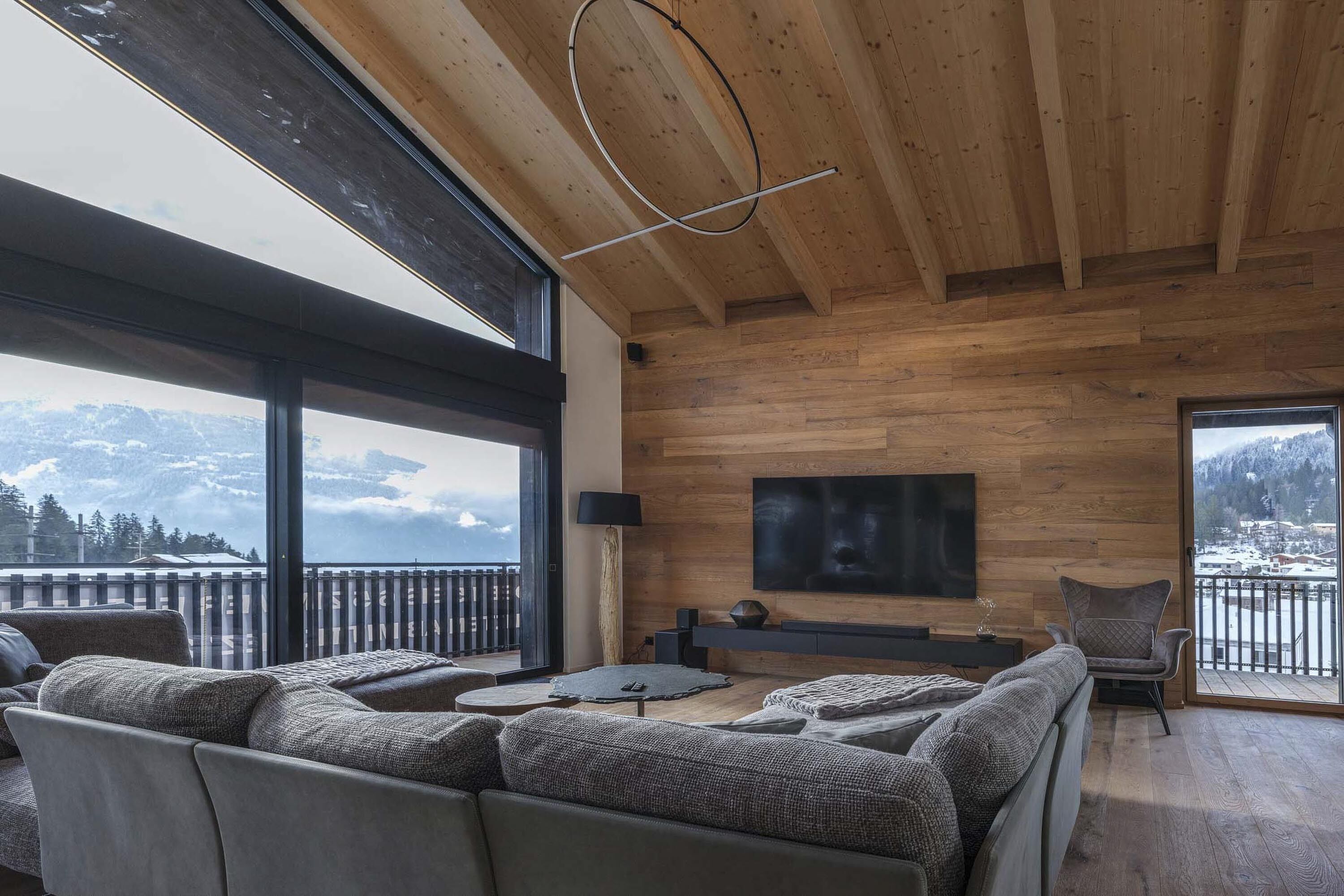 A spacious living room with a large sofa, a coffee table, and a TV mounted on the wall. The room has wooden paneling and large windows offering a view of mountains and a snowy landscape.