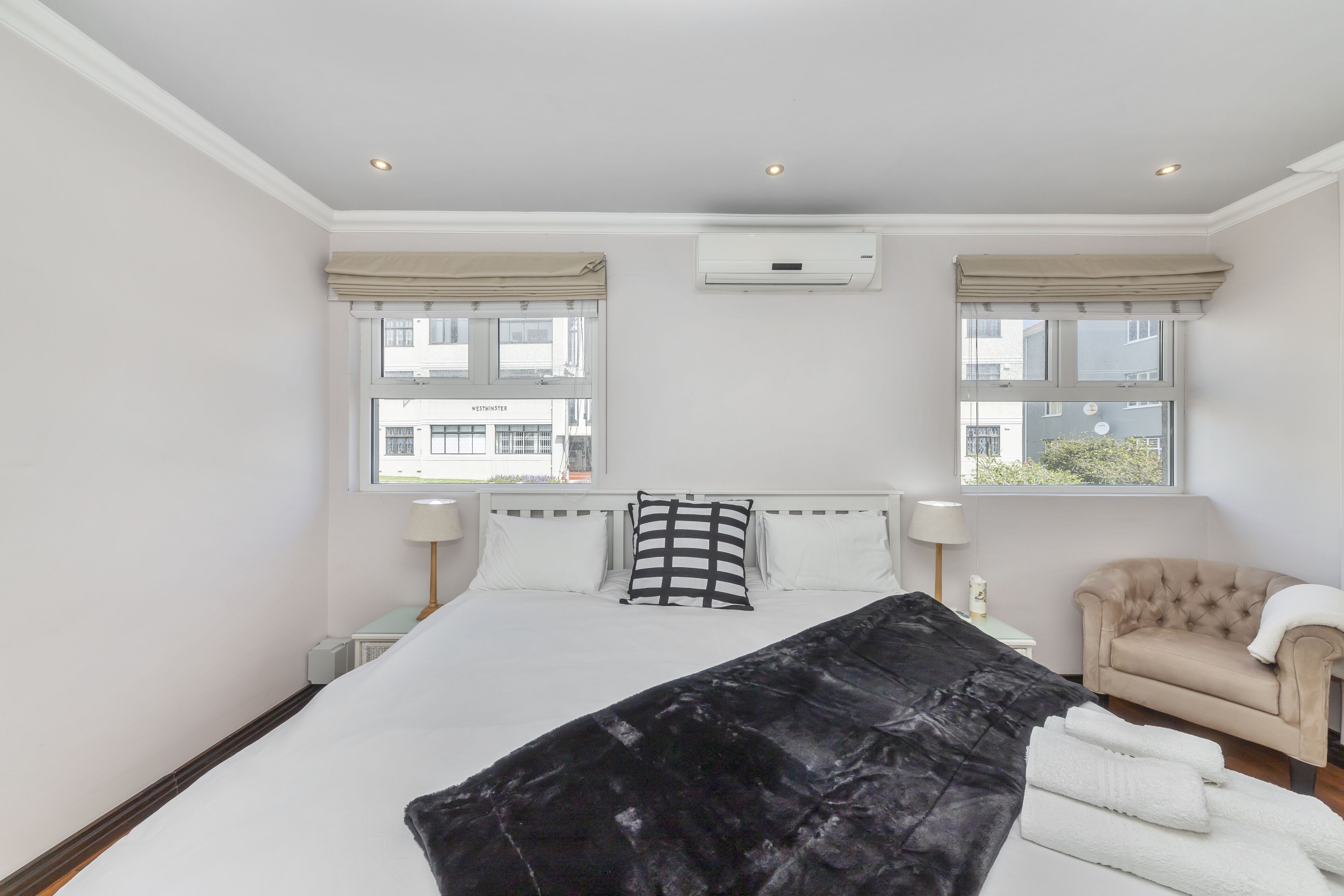 A hotel room with a large bed, two bedside tables, a chair, and an air conditioning unit.