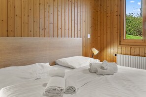 A neatly made bed with white linens and rolled towels, a bedside lamp, and a window with a view of greenery. - Logement Spacieux au Coeur du Val D'illiez (Val-d'Illiez)