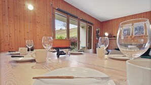 A dining table set with plates, cutlery, and wine glasses, with a view of a hillside through large windows.
