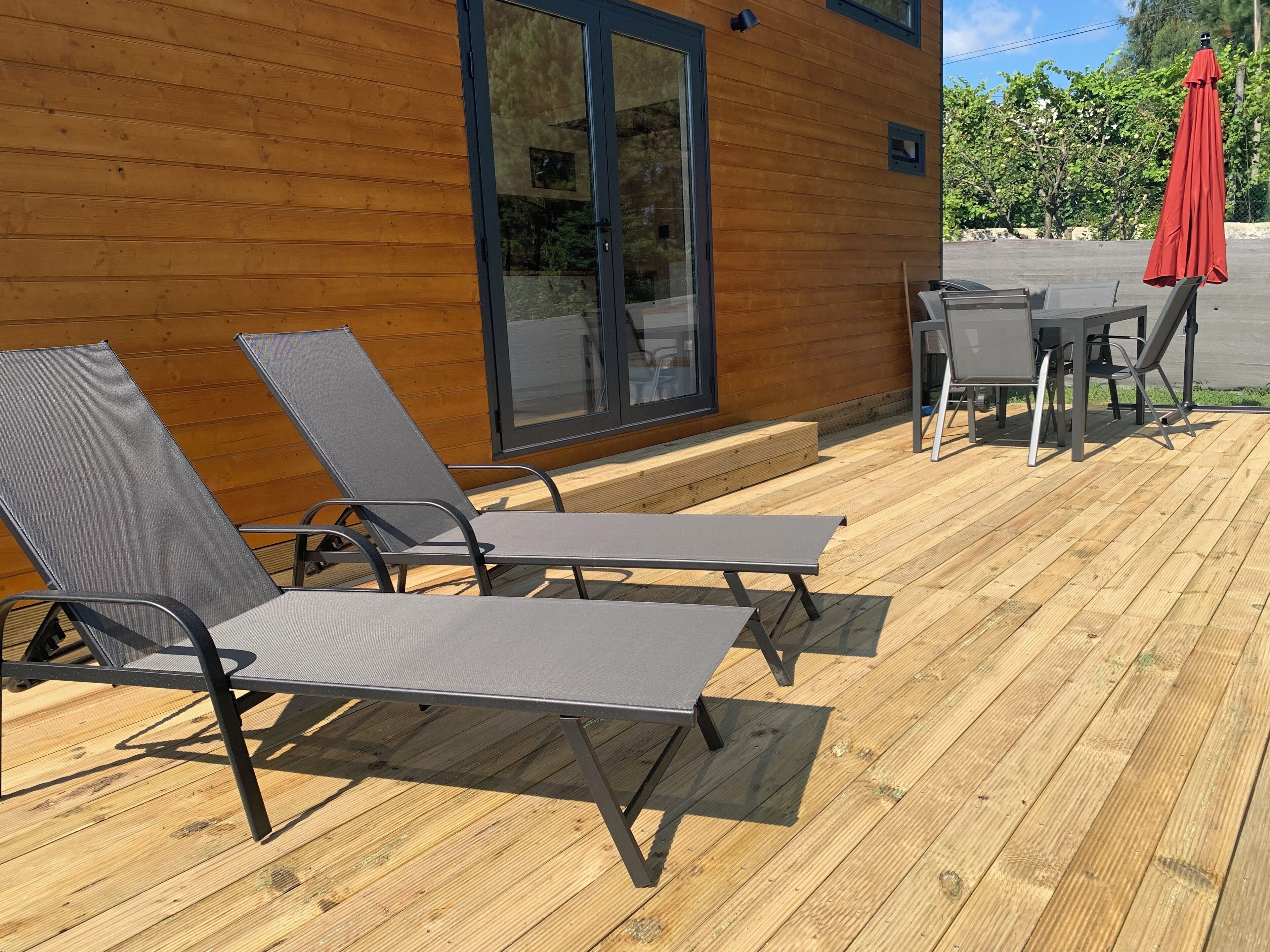 Une terrasse en bois avec deux chaises longues et une table, un parasol rouge et un bâtiment doté de grandes portes vitrées.