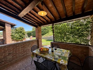 A covered patio area with a table set for dining, a hanging light fixture, and a view of a landscaped garden. - Agriturismo Case Graziani - Frantoio 2 (Citta della Pieve)