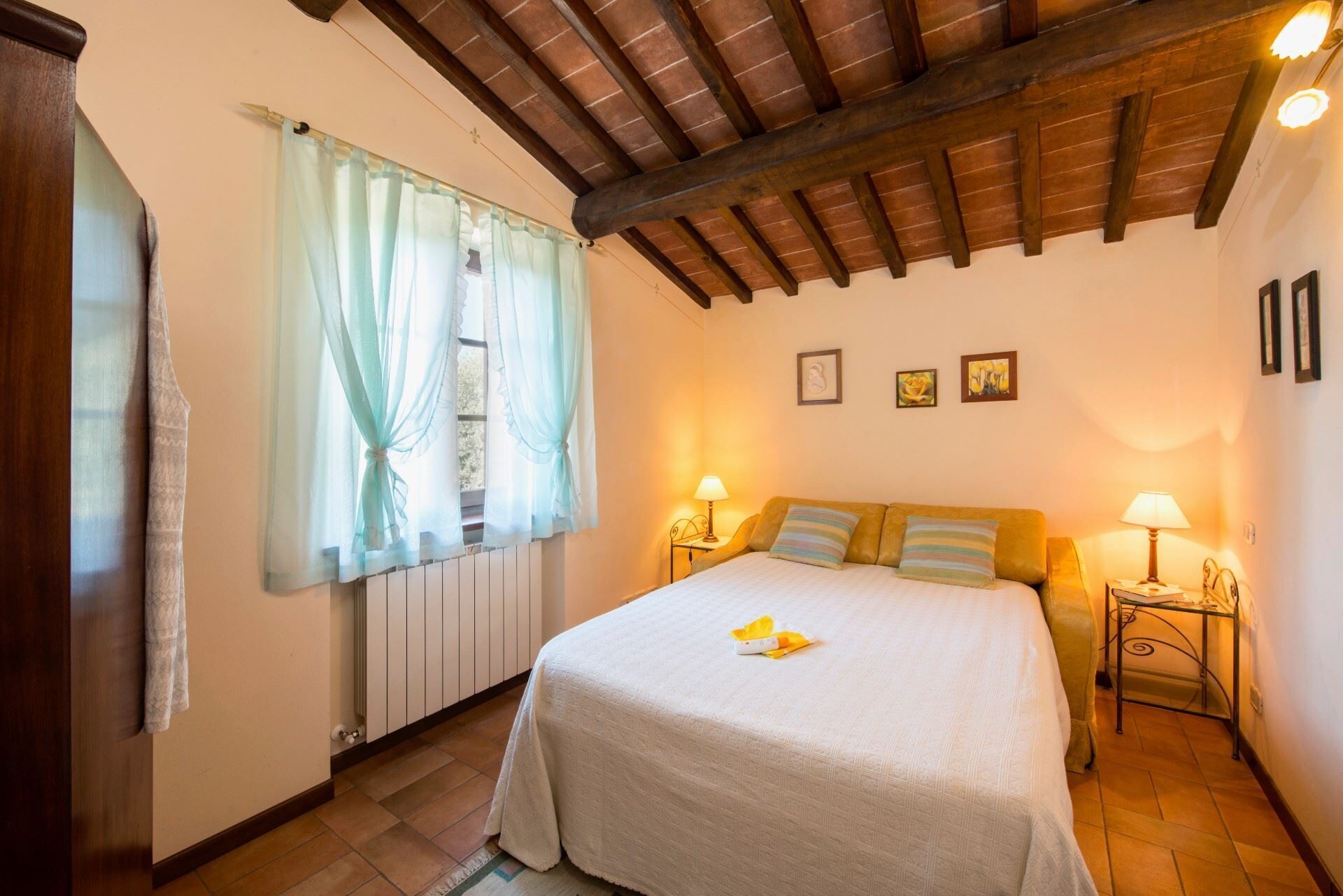 Un dormitori amb un llit, dues taules de nit amb làmpades, un radiador i una finestra amb cortines.