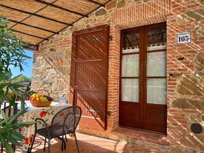 Un edificio de piedra con puerta y ventana de madera, una mesa con una cesta de fruta y una silla.