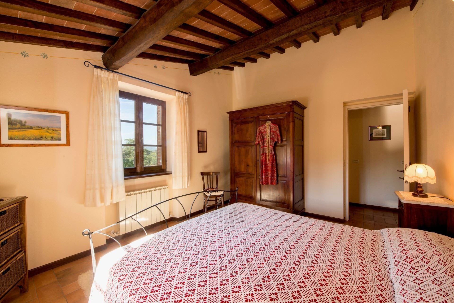 Una camera da letto con soffitto in legno, un letto con copriletto decorato, una finestra con tende, un armadio in legno e un comodino con una lampada.