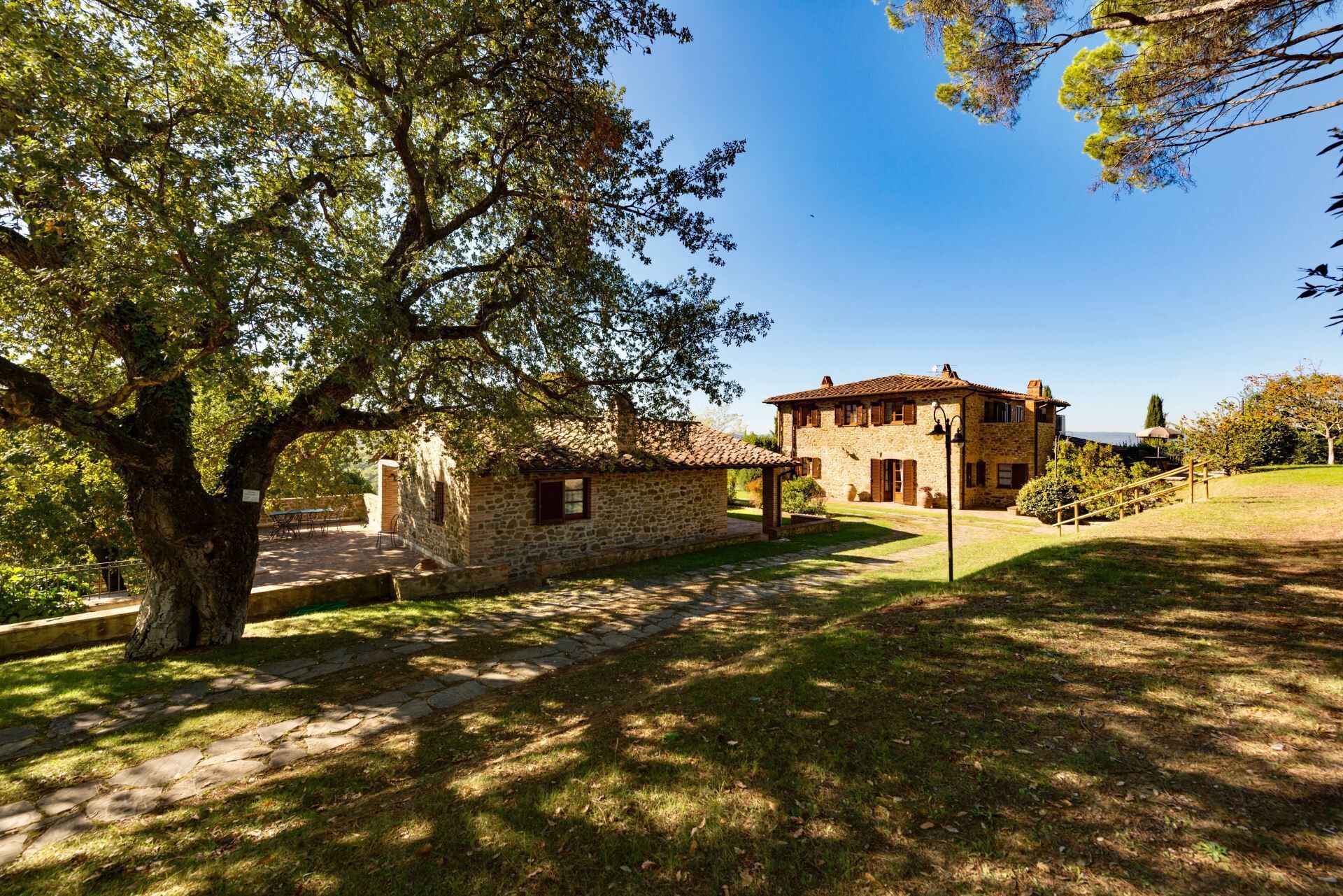 Una casa in pietra con tetto in tegole, circondata da un vialetto in pietra e un albero secolare.