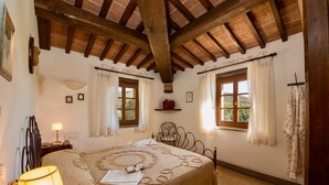 A bedroom with a wooden ceiling, two windows with curtains, a bed with a patterned bedspread, a bedside lamp, and a small shelf with a red bag. - Agriturismo Malagronda - Pino (Citta della Pieve)