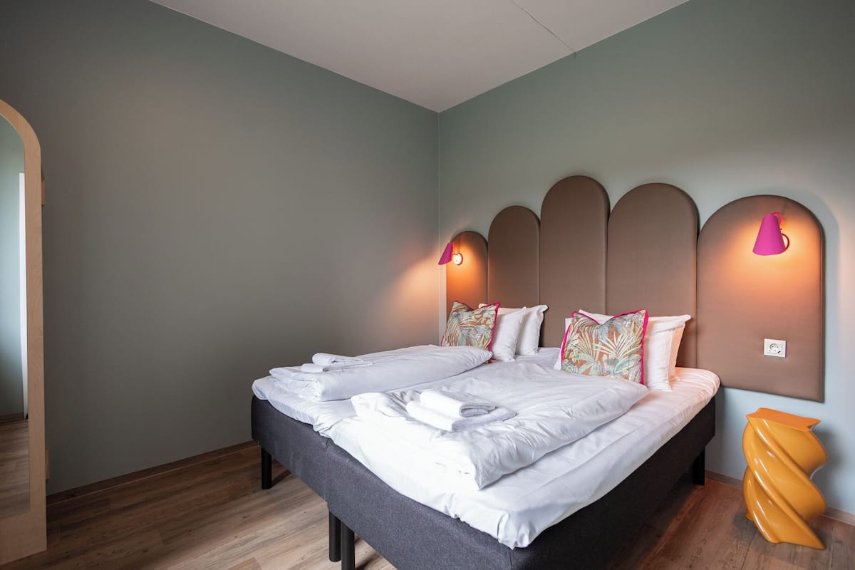 Una cama doble con sábanas blancas, suelo de madera y un llamativo detalle naranja.