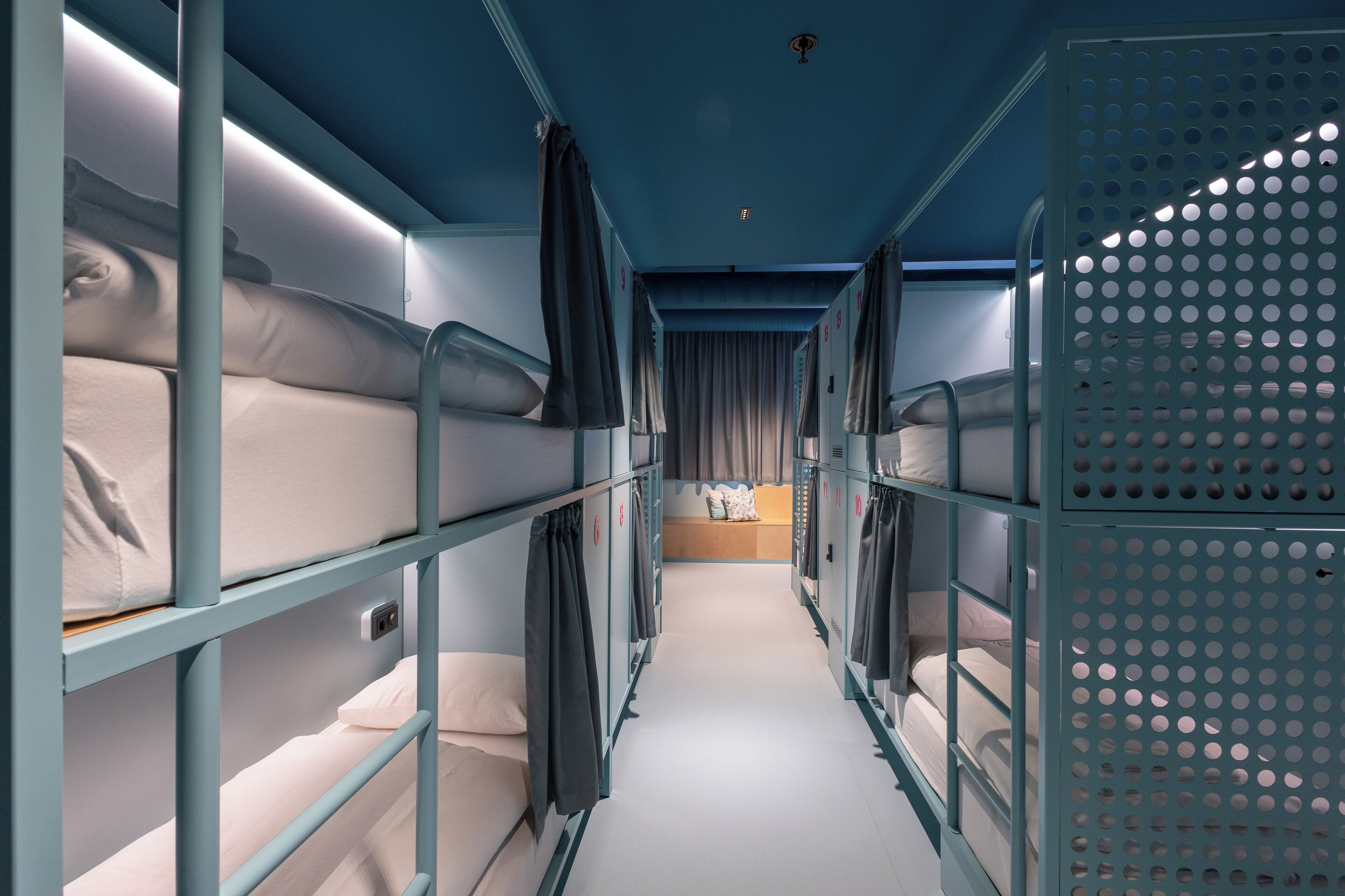 A narrow corridor with bunk beds on either side, a central walkway, and a perforated metal partition on the right.