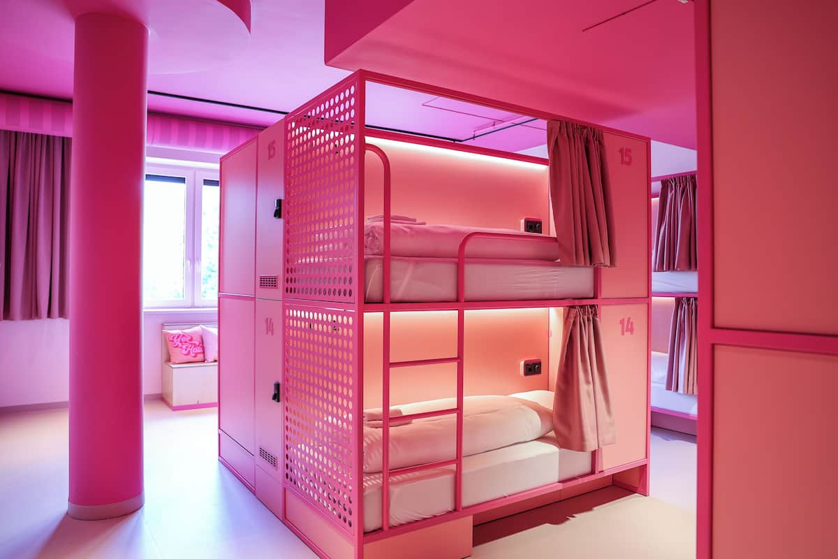 Una habitación con temática rosa, con literas, una ventana con cortinas y una pequeña zona de estar con cojín.