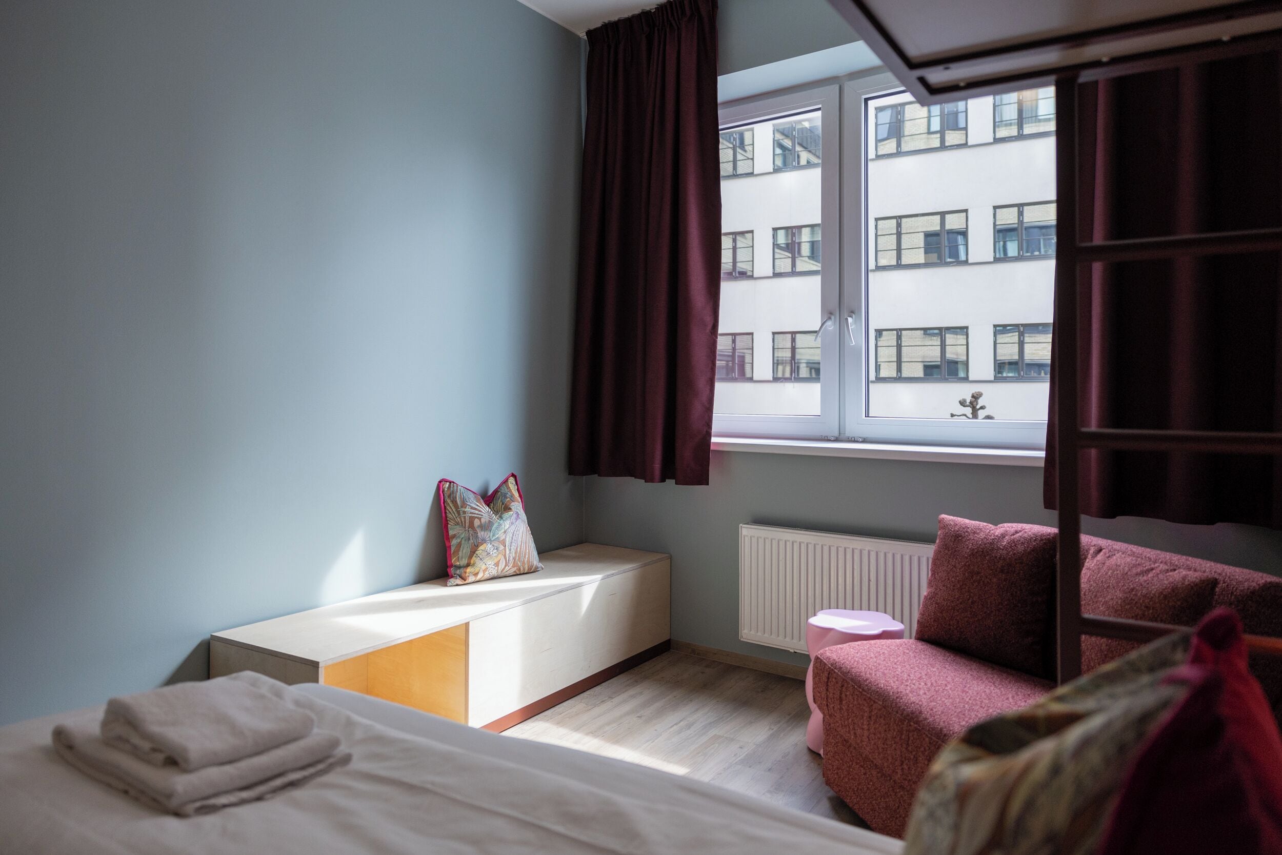 A bedroom with a bed, a bedside table, a window with curtains, a radiator, and a view of buildings outside.