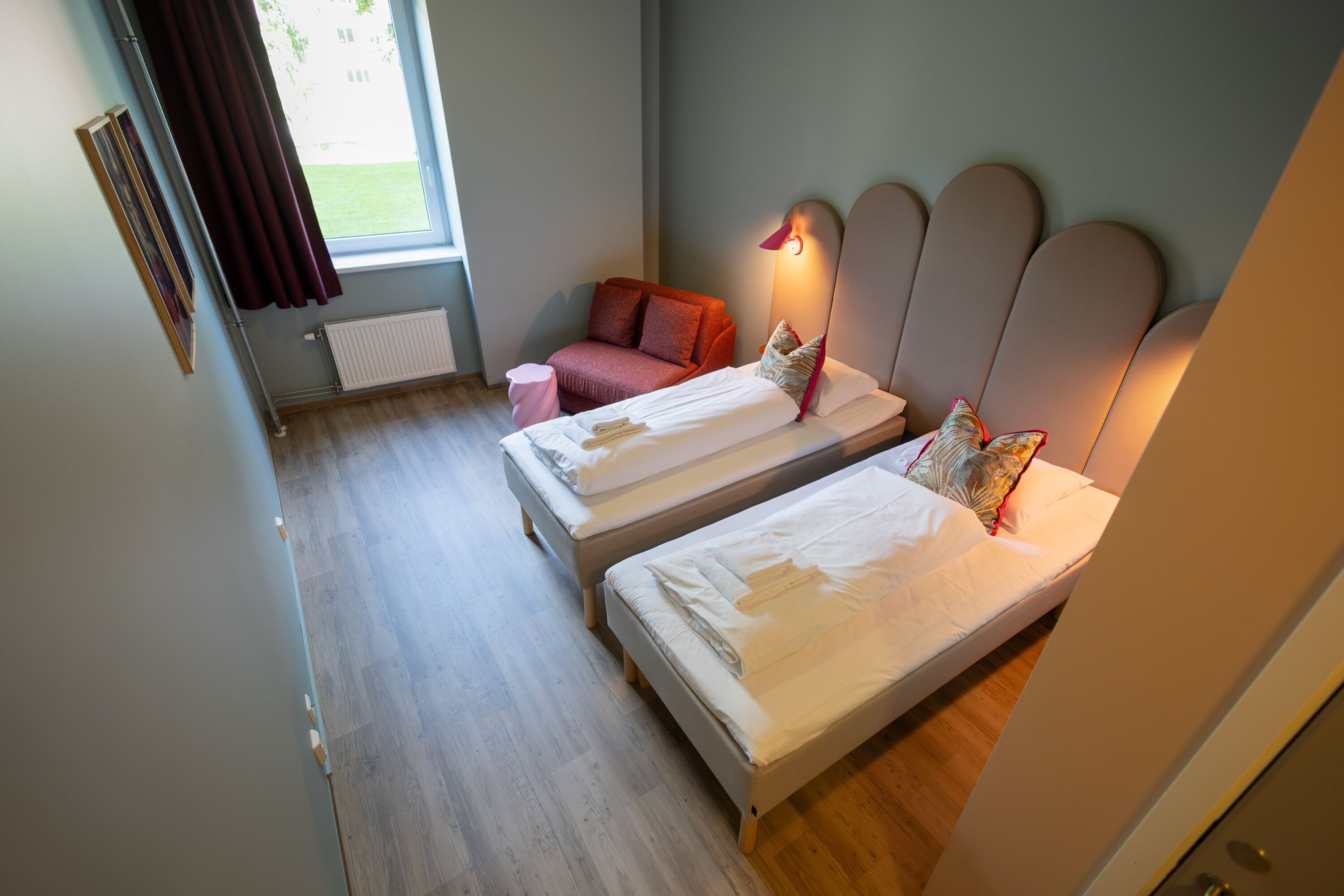 A bedroom with two beds, a radiator, a window with curtains, and a wall-mounted light.