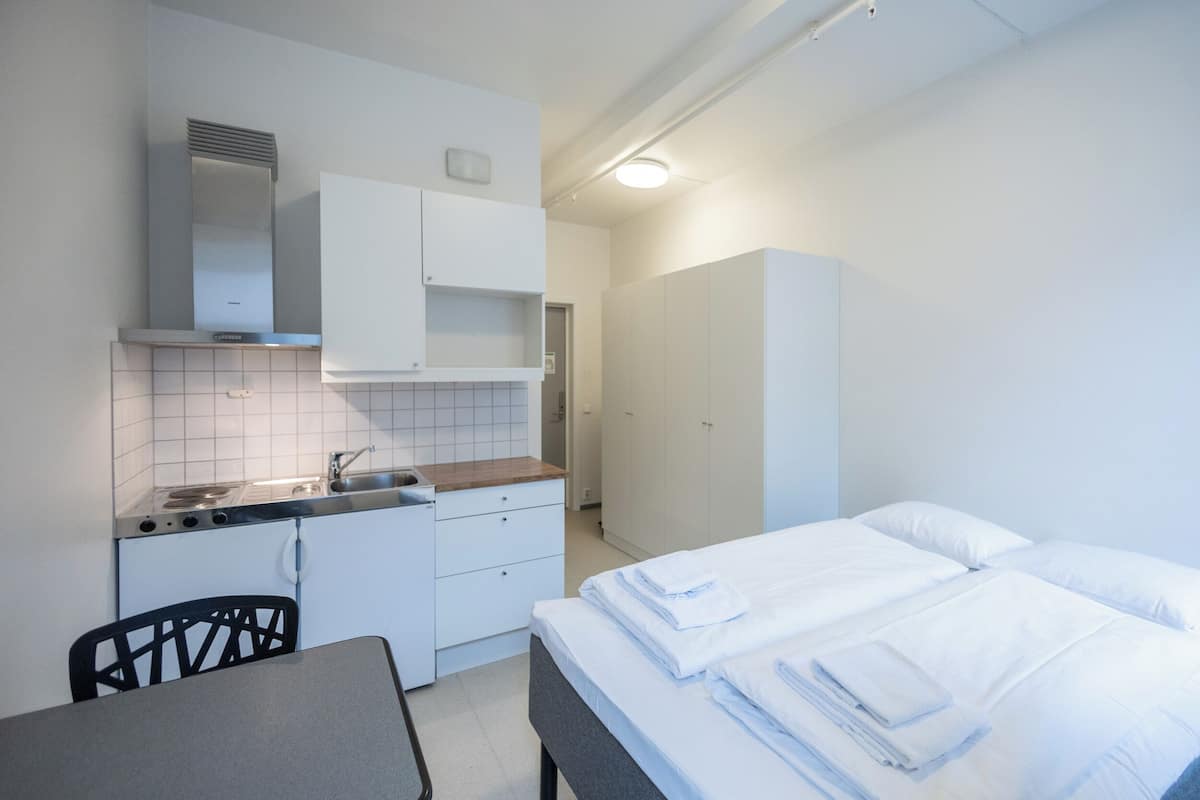 Una habitación pequeña con kitchenette, una cama con sábanas blancas impecables y una silla.
