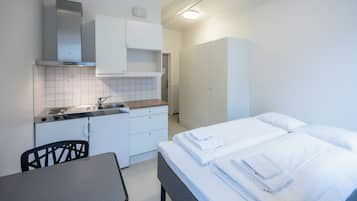 Una habitación pequeña con kitchenette, una cama con sábanas blancas impecables y una silla.