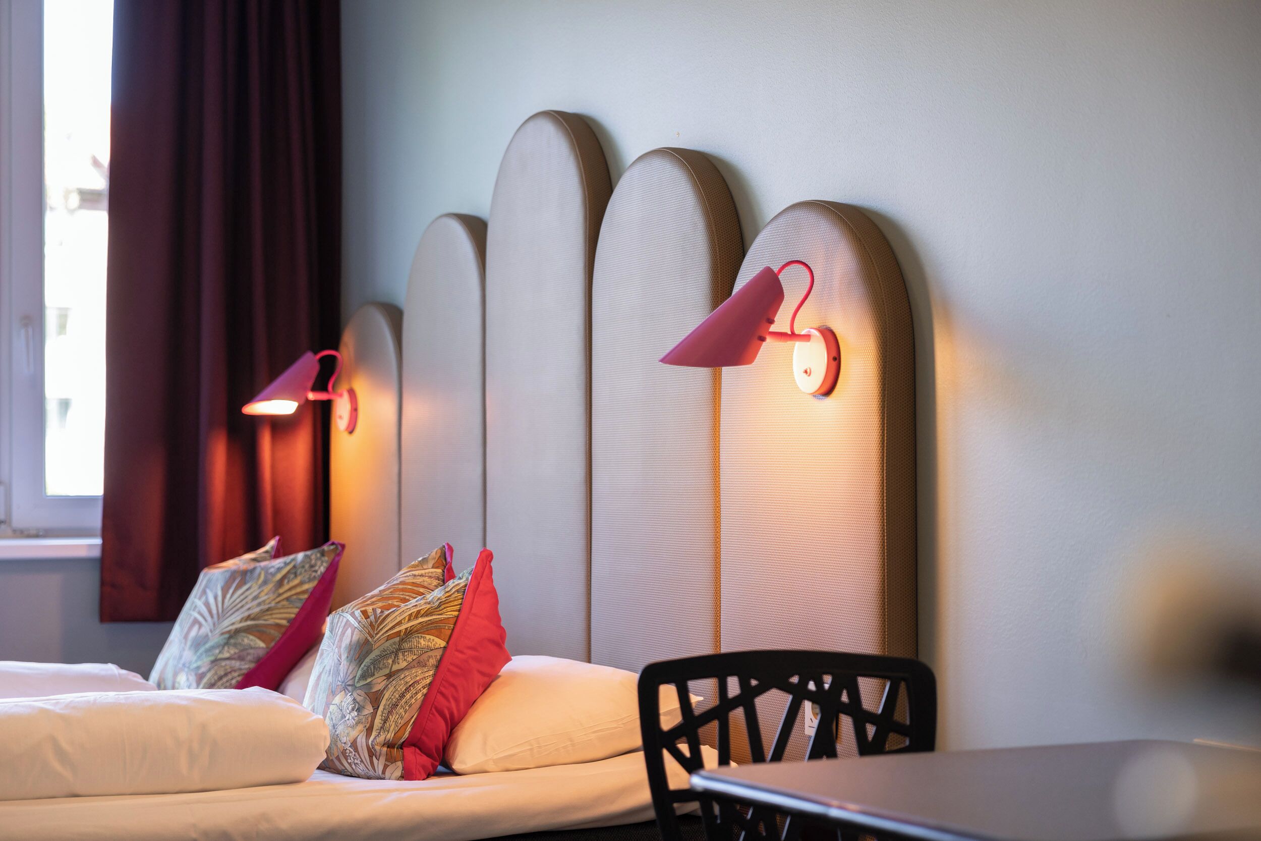 A hotel room with a bed, a table, and a chair. There are decorative pillows on the bed and a wall-mounted light fixture.